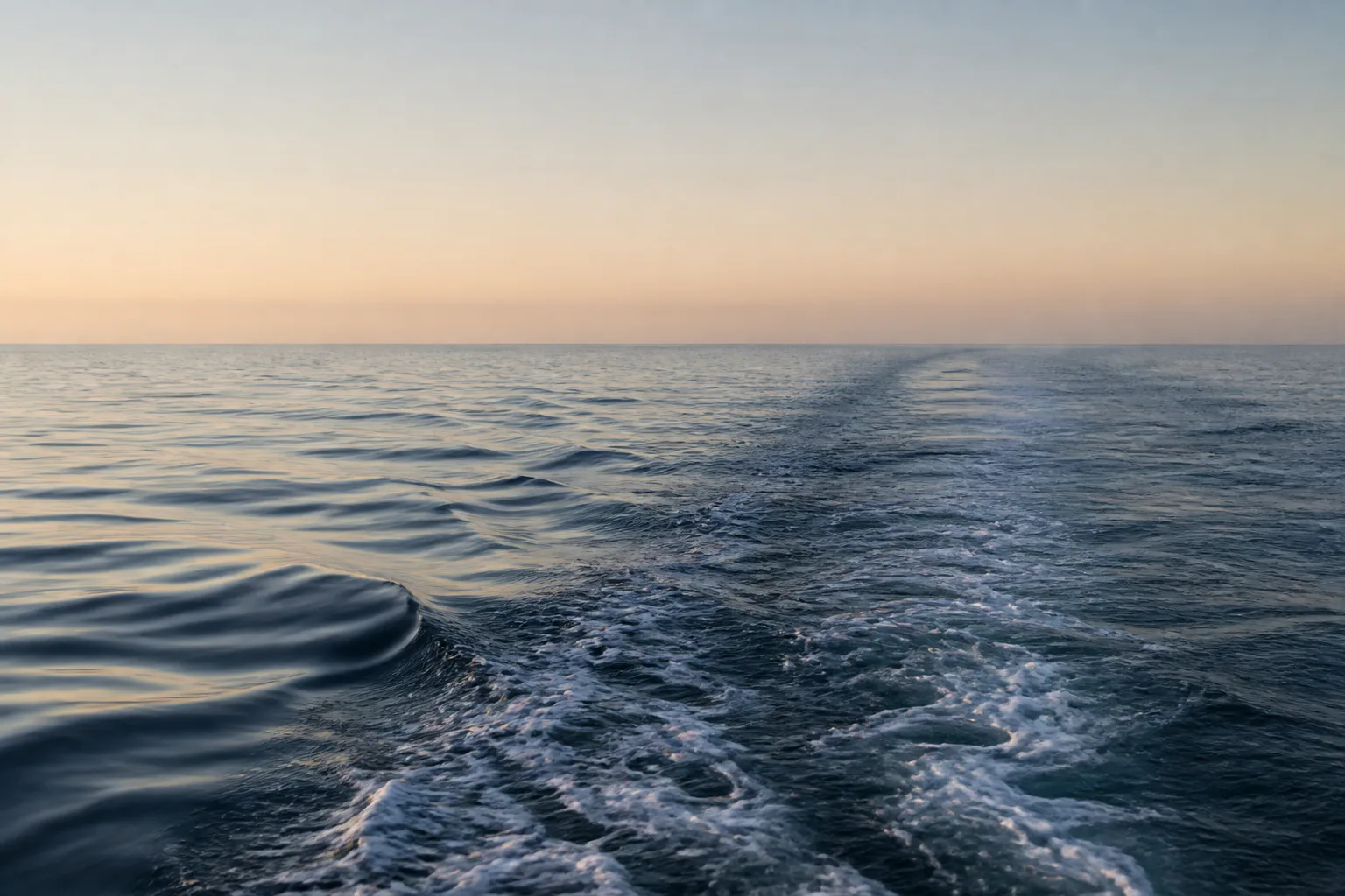 A wake recedes towards a calm sea horizon at dawn.