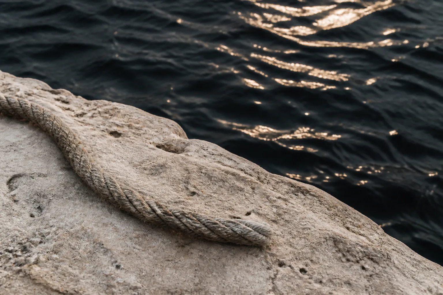 Rope on pale stone beside dark water lit by the sun.